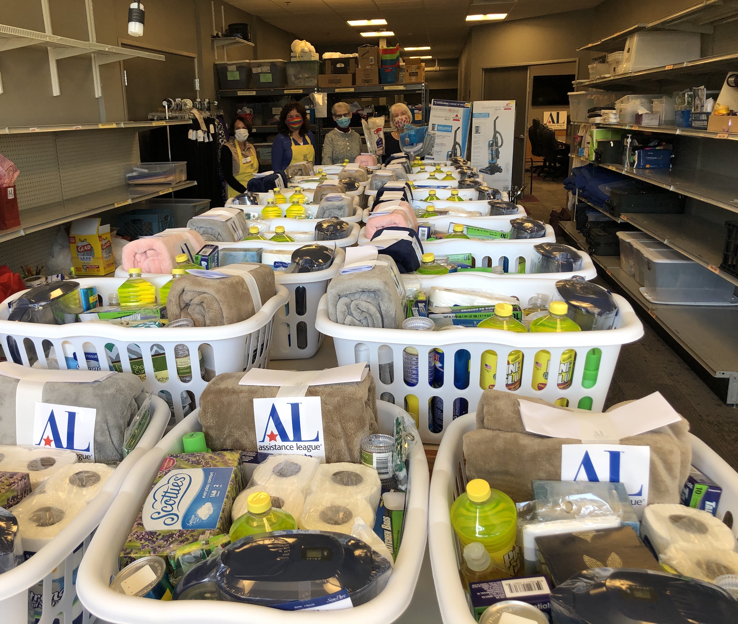 A long table covered in Fresh Start baskets filled with toiletries and cleaning supplies. Four volunteers pose at the end of the table in the background.